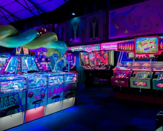 An indoor arcade filled with vibrant gaming machines lit with neon blue and pink lights. The scene includes various arcade games with flashing screens and colorful displays, and a large overhead structure resembling a green snake. The background shows illuminated signs and themed decor, contributing to an immersive entertainment atmosphere.