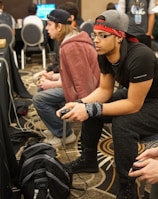 Several people are sitting in a room with gaming consoles and monitors, focusing intently on their screens. The setting appears to be a casual gaming event, with participants holding controllers while playing video games. The room has a casual atmosphere with chairs and tables set up for gaming.