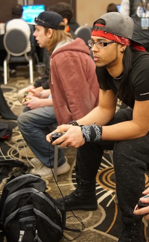 Several people are sitting in a room with gaming consoles and monitors, focusing intently on their screens. The setting appears to be a casual gaming event, with participants holding controllers while playing video games. The room has a casual atmosphere with chairs and tables set up for gaming.