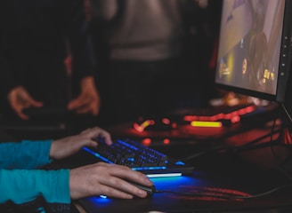 A person is engaged in a video game at a computer setup, using a keyboard and mouse. The scene is dimly lit, with the glow from the monitor reflecting off the hands of the player. Several other people are present in the background, indicating a social or competitive gaming environment.