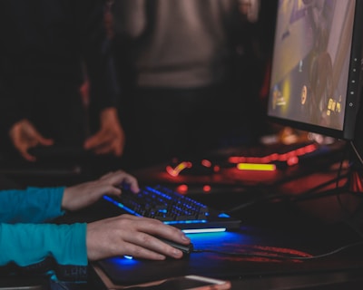 A person is engaged in a video game at a computer setup, using a keyboard and mouse. The scene is dimly lit, with the glow from the monitor reflecting off the hands of the player. Several other people are present in the background, indicating a social or competitive gaming environment.