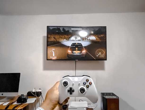 A person is holding a gaming controller in front of a flat-screen television displaying a racing video game. The room has a computer setup on a wooden desk with cables, a desktop computer monitor, and other accessories. The focus is on the interaction between the person and the gaming environment.