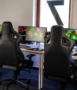 A row of gaming stations equipped with large monitors, gaming chairs, keyboards, and headphones. The setup is organized and ready for gaming, featuring a colorful screen display. The room has a modern and tech-savvy ambiance.