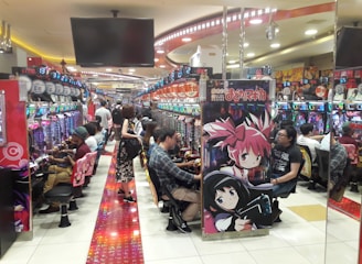 A bustling arcade filled with people playing various gaming machines. Brightly colored displays and animated characters are prominent on posters and screens. The atmosphere is lively with patrons seated in rows, focusing on the games. Overhead signage and electronic scoreboards are visible, contributing to the busy and vibrant environment.