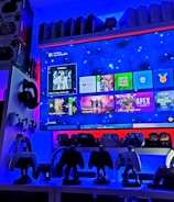 A modern gaming setup featuring a large wall-mounted screen displaying a gaming console interface. The room is decorated with gaming-themed items such as controllers and collectibles. The ambient lighting includes shades of blue and red, creating a vibrant atmosphere.