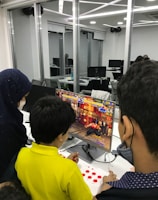 A group of children is gathered around a desktop computer, engaged in playing a colorful, action-packed arcade game displayed on the monitor. The setting appears to be a modern, well-lit office or gaming space with large glass windows, showing multiple computer workstations in the background.