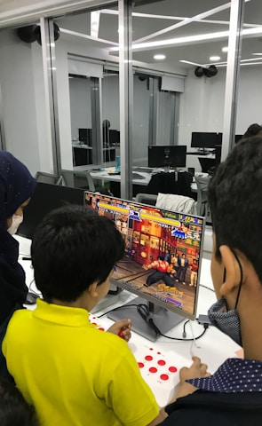 A group of children is gathered around a desktop computer, engaged in playing a colorful, action-packed arcade game displayed on the monitor. The setting appears to be a modern, well-lit office or gaming space with large glass windows, showing multiple computer workstations in the background.