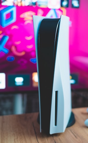 A gaming console is placed on a wooden table with a game controller nearby. In the background, a television screen displays colorful graphics, including geometric shapes and vibrant colors.